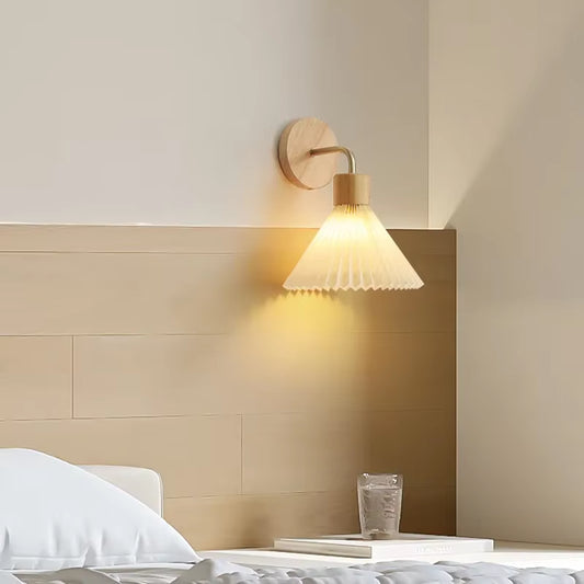 Pleated cone wall sconce on a light wood plate, lit, above a bed.