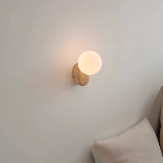 Wall sconce with a white glass globe on a light wood base, lit, seen from the side above a cushion.