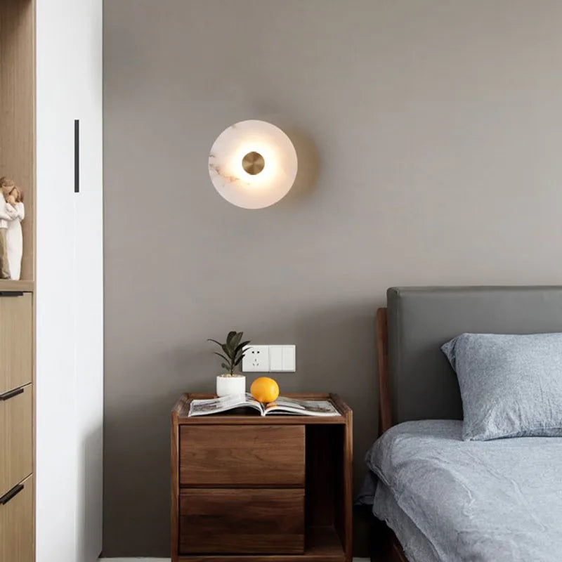 Round marble disc wall sconce lit above a wood nightstand beside a bed.