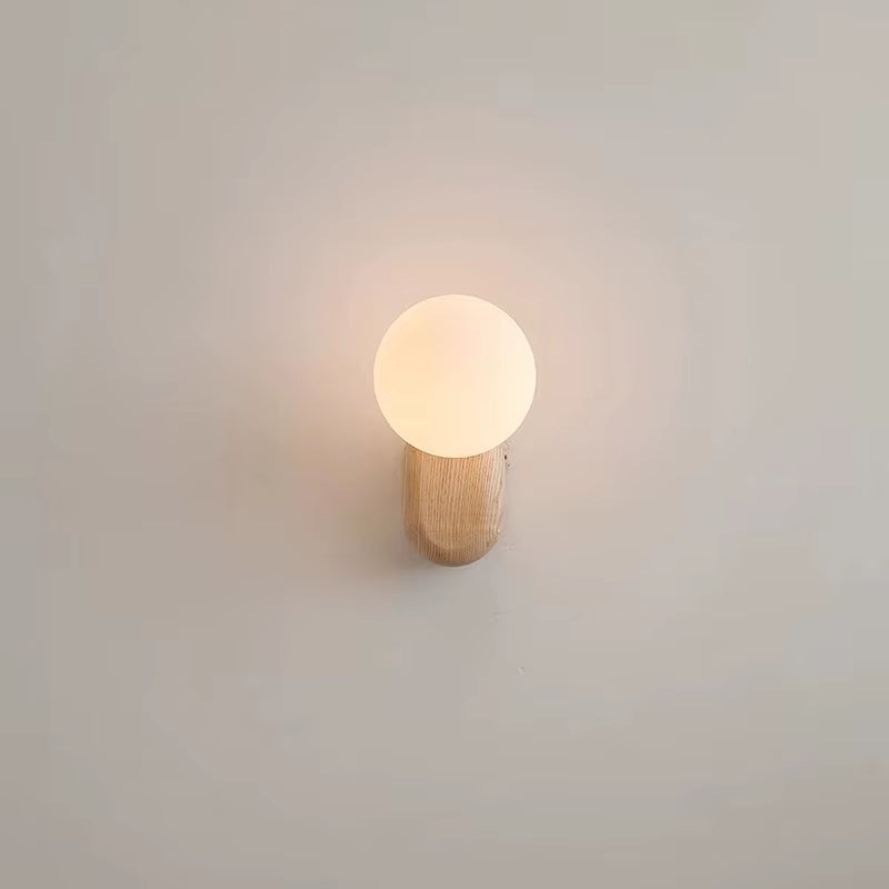 Wall sconce with a white glass globe on a light wood base, lit, seen from the front on a pale wall.