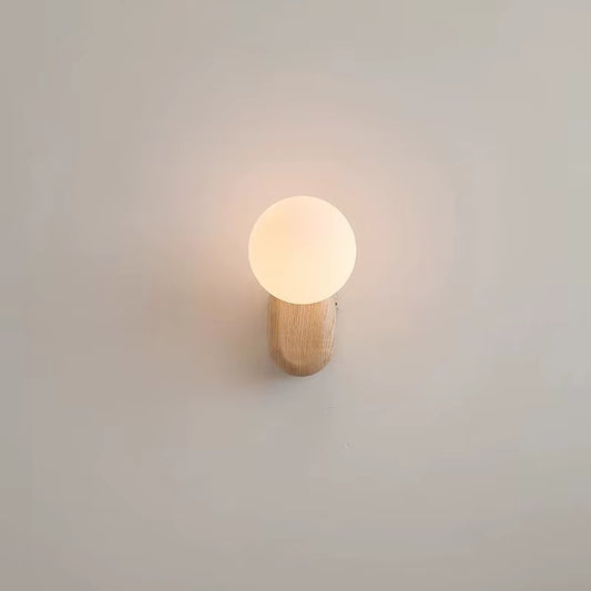 Wall sconce with a white glass globe on a light wood base, lit, seen from the front on a pale wall.