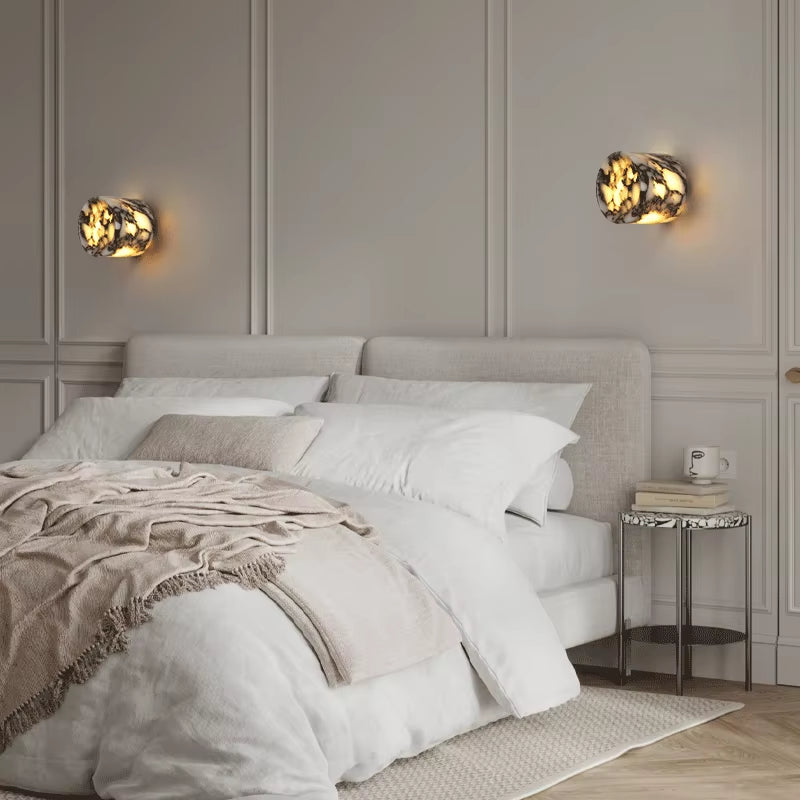 A pair of marble cylinder wall sconces, lit, on a panelled wall above a bed.
