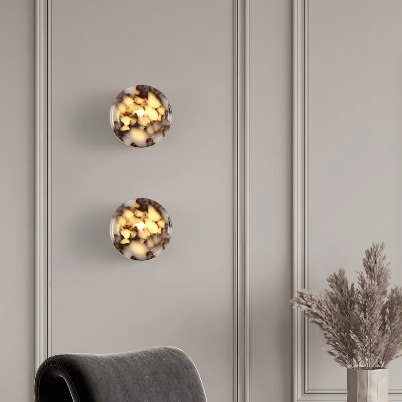 Two marble cylinder wall sconces seen from the front, lit, on a panelled wall.