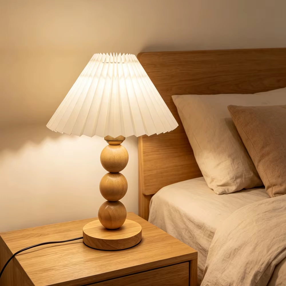 Natural wood stacked-sphere table lamp with a white pleated shade, lit, on a nightstand beside a bed.