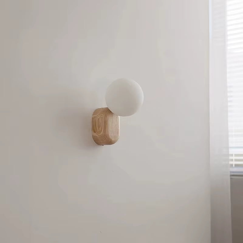 Wall sconce with a white glass globe on a light wood base, unlit, on a wall beside a window.