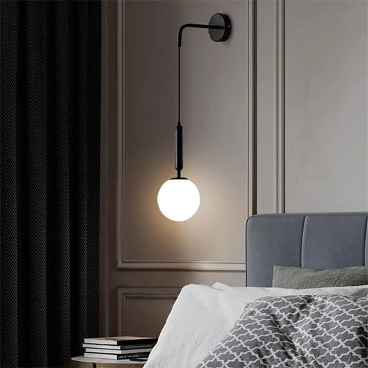 Frosted glass globe wall sconce on a long black metal drop arm, lit, beside a bed and a dark curtain.