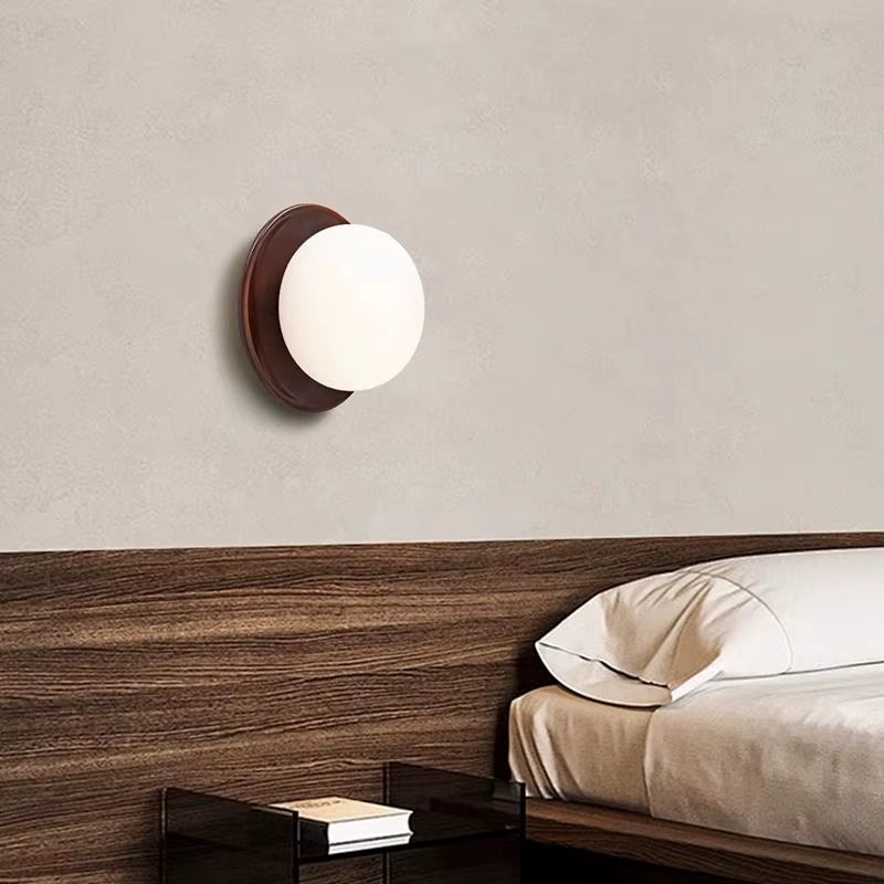 Walnut-tone wood disc wall sconce with a frosted glass globe above a wood headboard.