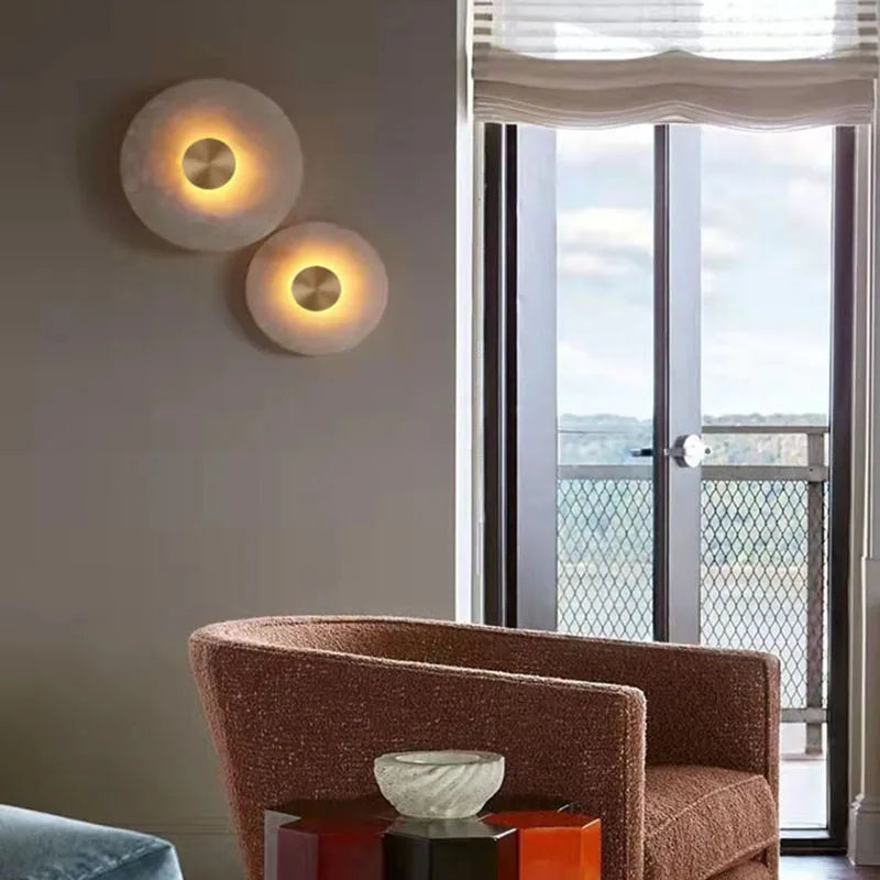 Two marble disc wall sconces lit on a taupe wall beside a window and an armchair.