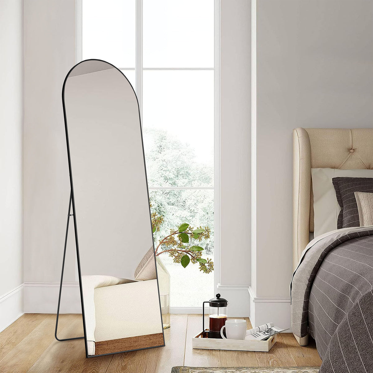 Arched full-length floor mirror with a thin black frame, standing on its stand beside a bed and a tall window.