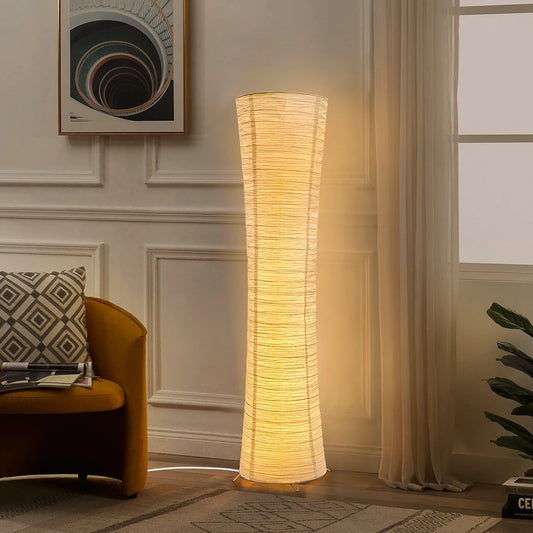 Rice paper column floor lamp glowing warm white beside an armchair in a paneled living room.