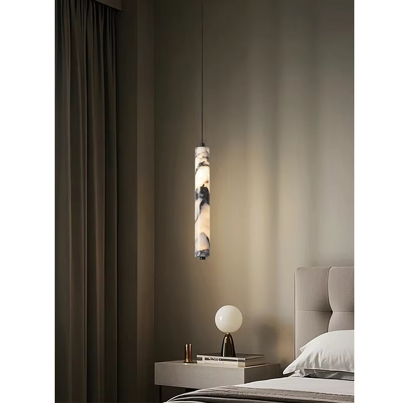 Slim marble cylinder pendant light, lit, hanging above a nightstand beside a dark curtain.