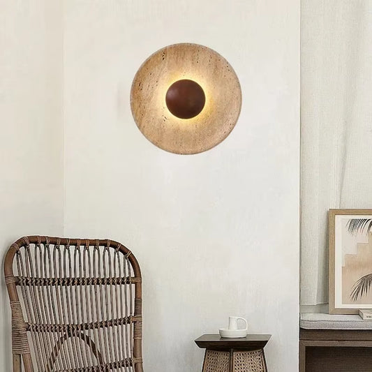 Round stone disc wall sconce with a dark wood center, lit, on a white wall above a rattan chair.