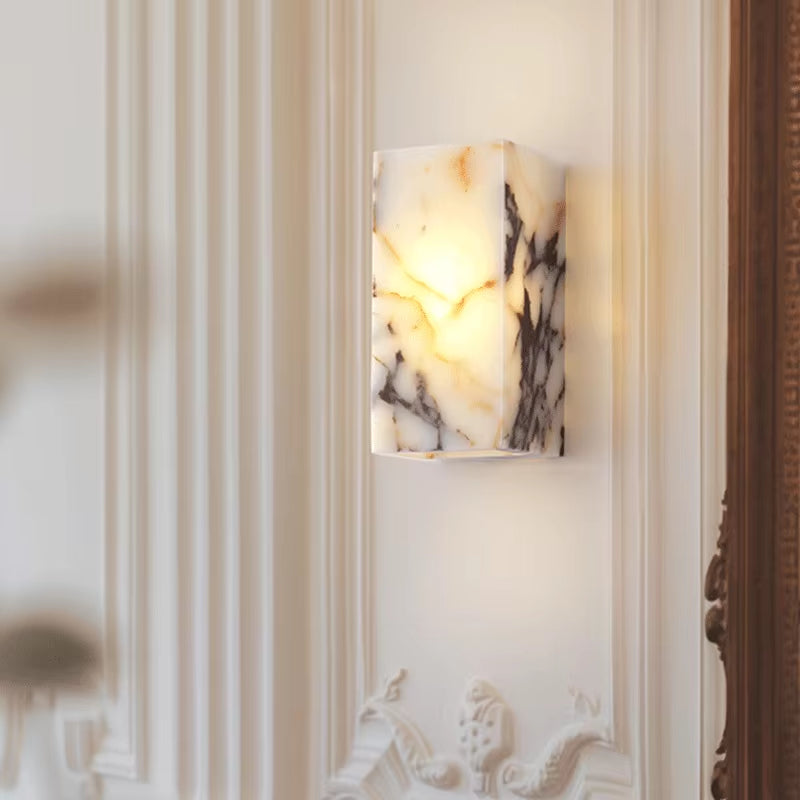 Stone block wall sconce, lit, on a panelled wall beside a carved wood mirror frame.
