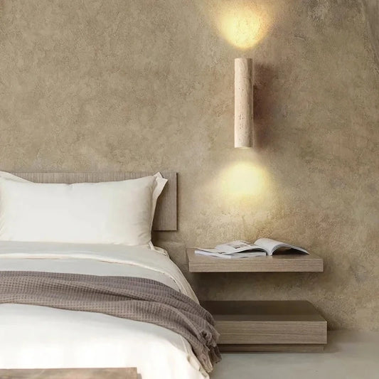 Travertine cylinder wall sconce on a walnut bracket lighting the wall above a bedside shelf.