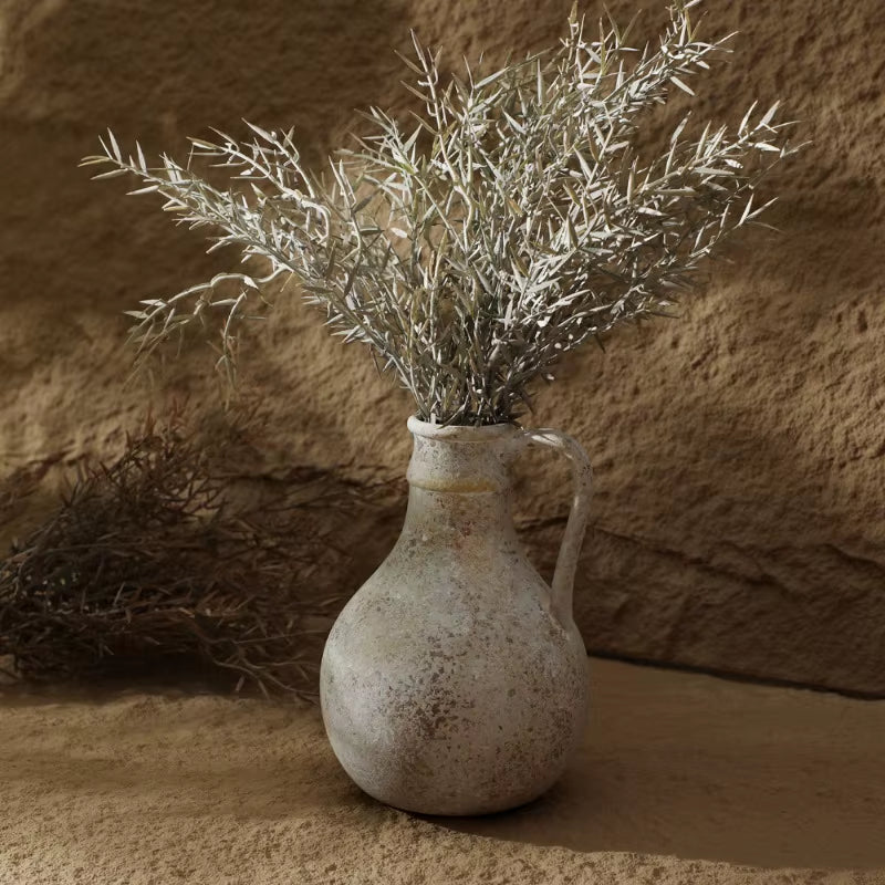 Round ceramic jug vase holding dried branches against a textured wall.