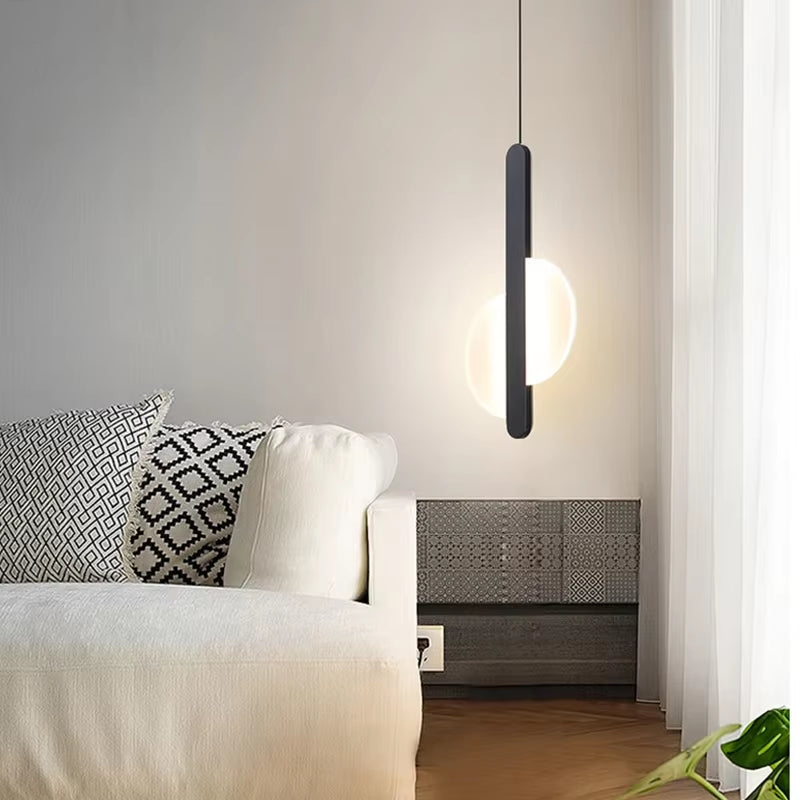Black bar pendant light with a split round disc, lit, hanging beside a white sofa.