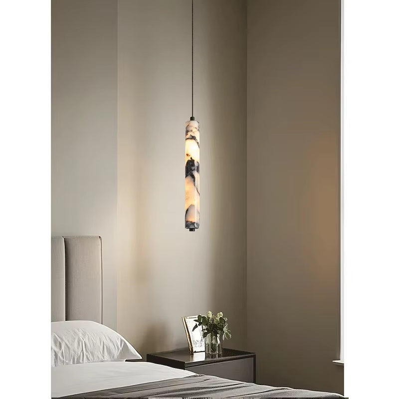 Slim marble cylinder pendant light, lit, hanging beside a bed above a dark nightstand.