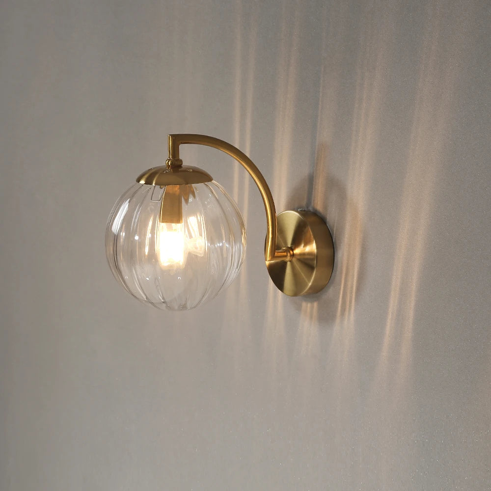 Glass wall sconce with a clear ribbed glass globe, lit, on a gold-color curved arm.