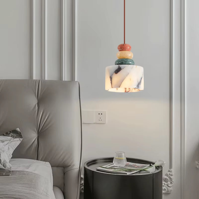 Stone drum pendant light, lit, hanging above a round nightstand beside a bed.