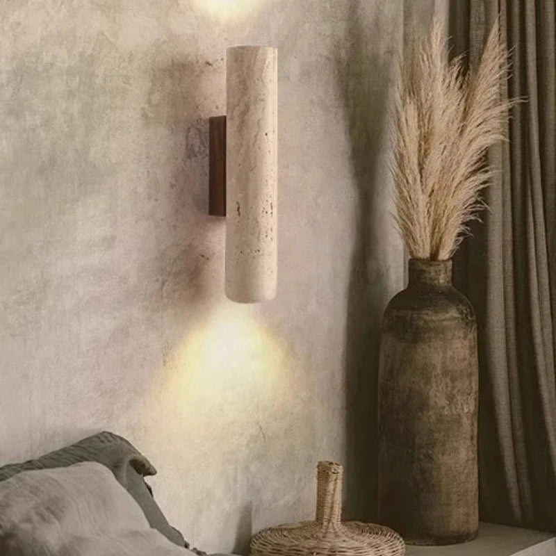 Travertine cylinder wall sconce casting light up and down on a plaster wall beside a vase.