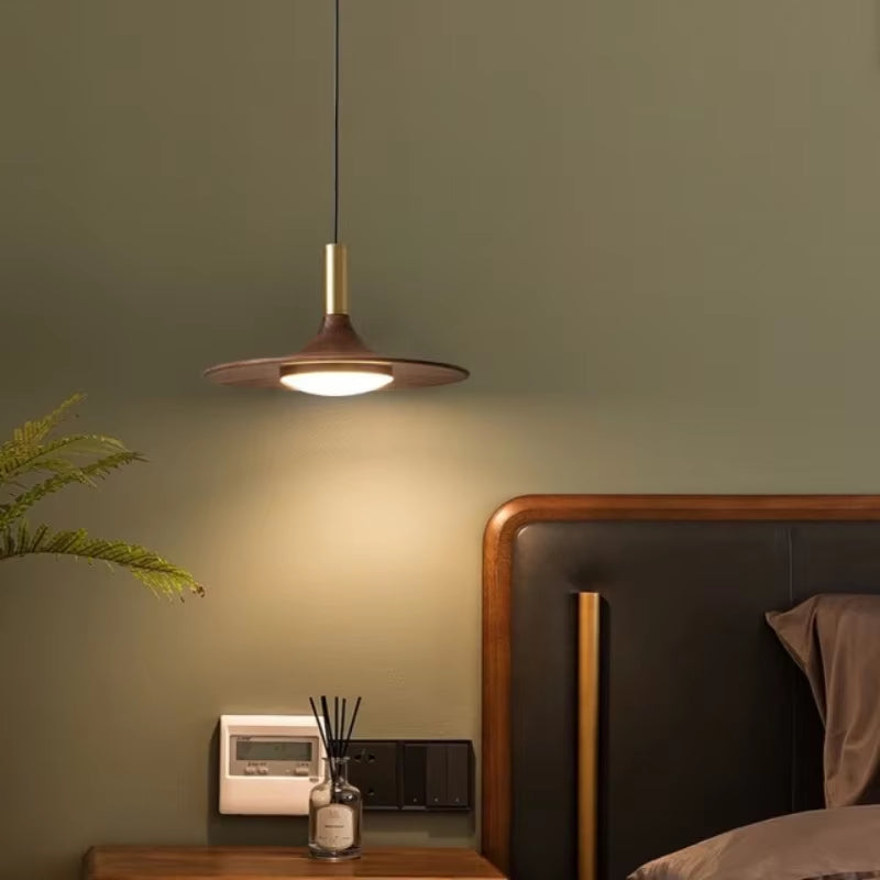 Flying saucer pendant light in a walnut-grain finish hanging beside a bed, lit.