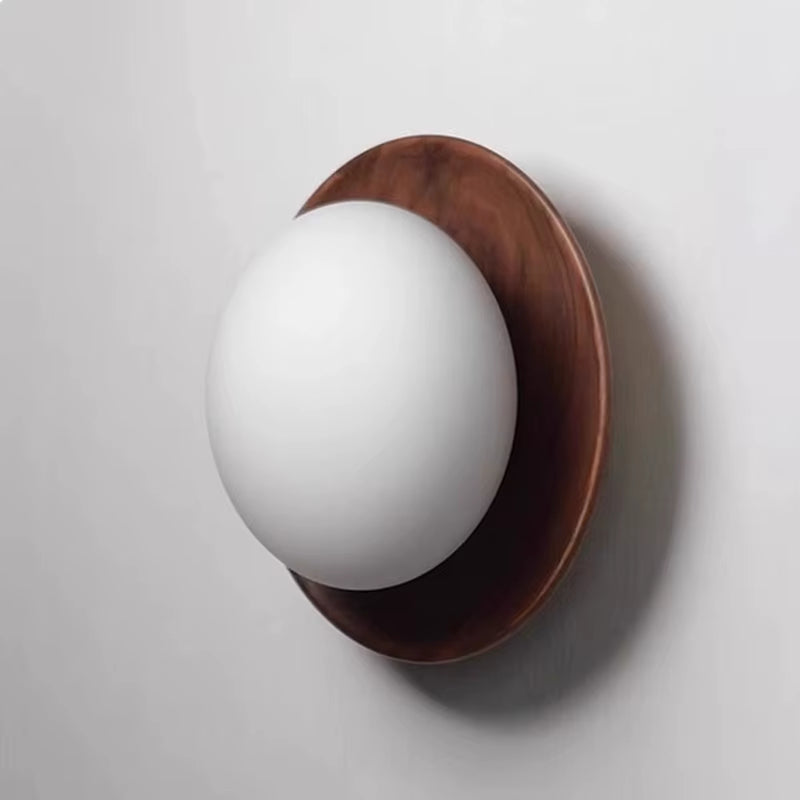 Wall sconce with a frosted glass globe on a walnut-tone wood disc, close view on a white wall.