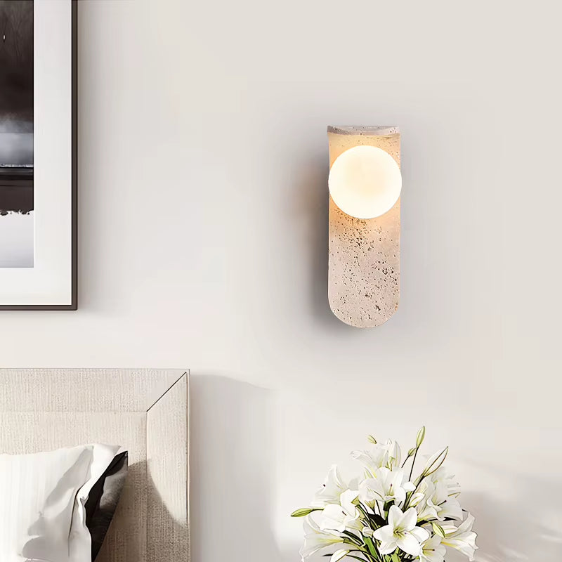 Travertine globe wall sconce lit on a bedroom wall beside a framed print