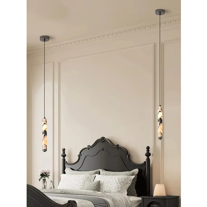 Two slim marble cylinder pendant lights, lit, hanging on either side of a bed with a dark headboard.