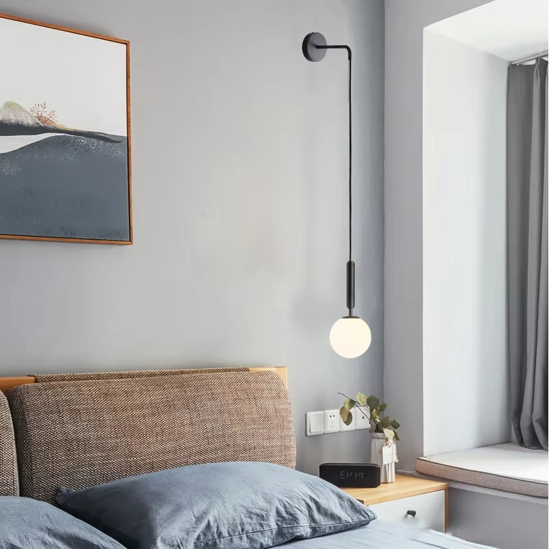 Frosted globe wall sconce on a long black metal drop arm beside a bed with a framed print.