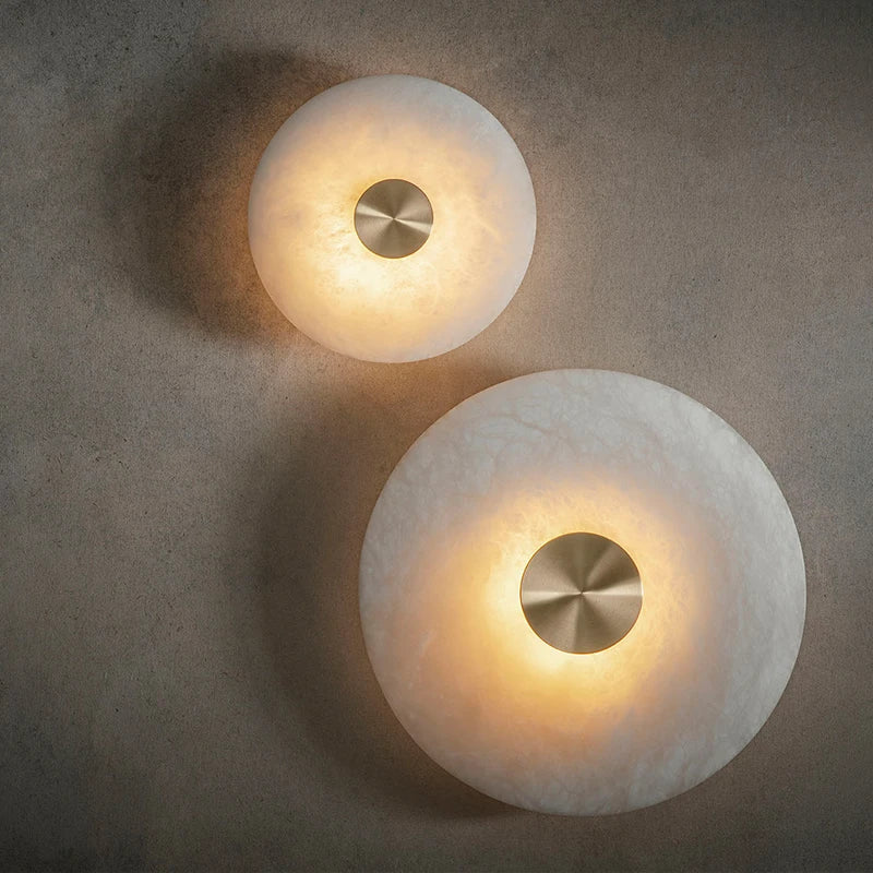 Two round marble disc wall sconces in two sizes with gold-color metal centers, lit, on a textured grey wall.