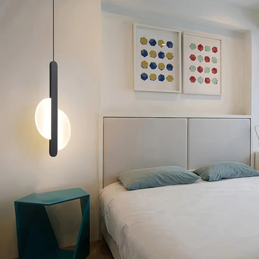 Black bar pendant light with a split round disc, lit, hanging beside a bed above a blue side table.