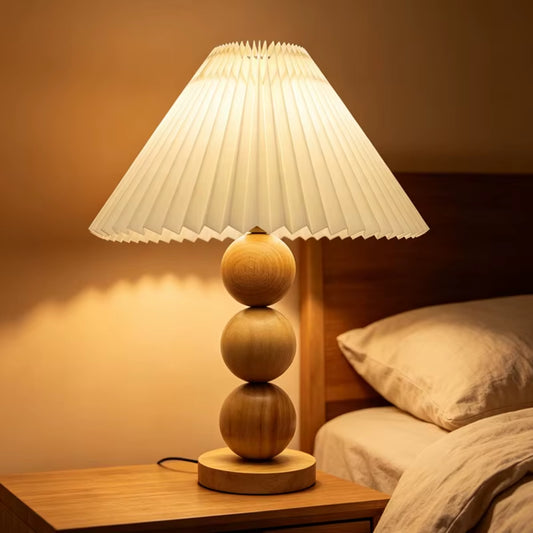 Natural wood stacked-sphere table lamp with a white pleated shade, lit, on a bedside table.