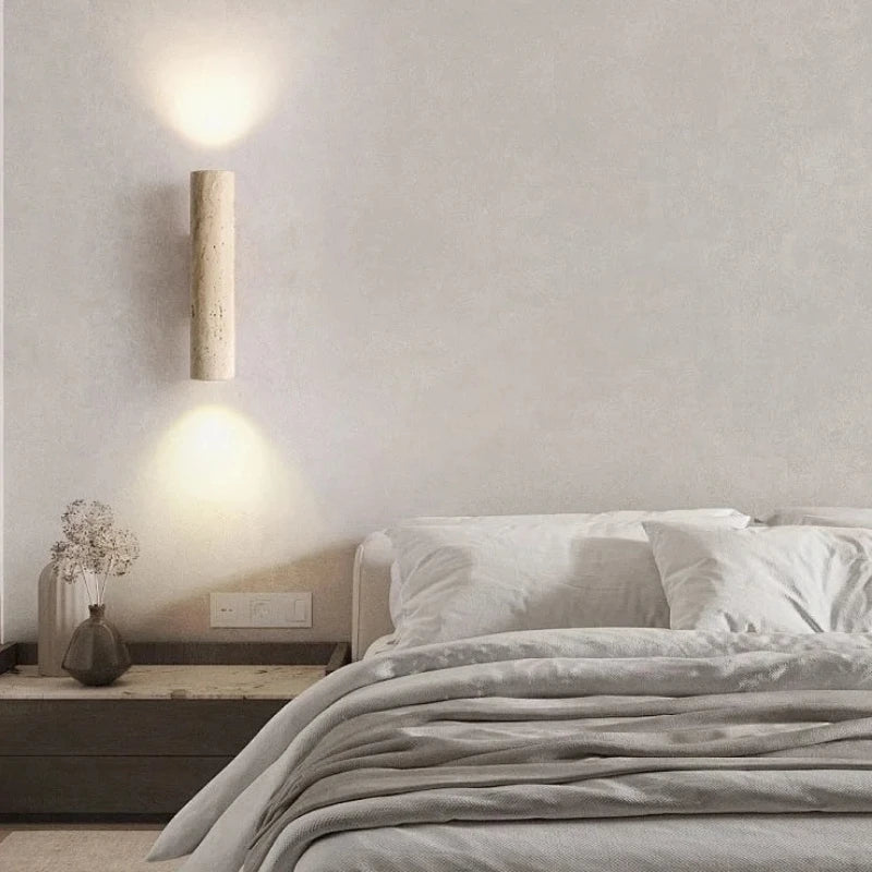 Travertine cylinder wall sconce lit above a bed with white linens.