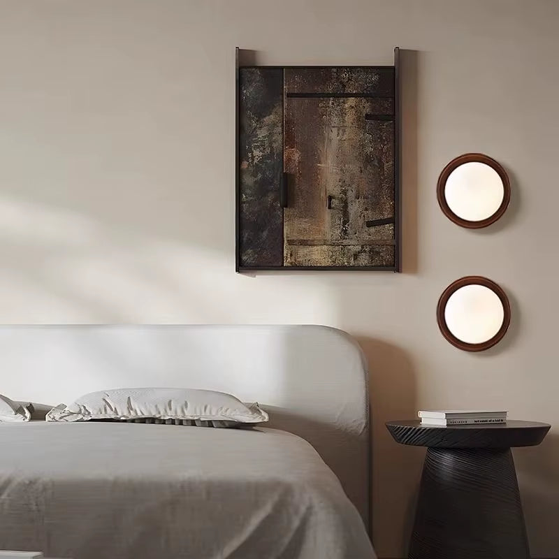Two walnut-tone disc wall sconces with frosted glass globes beside a bed and a framed artwork.