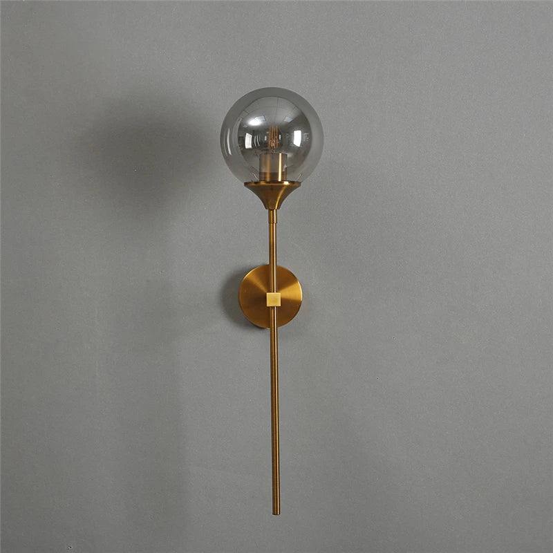 Smoke gray glass globe wall sconce on a long gold-color metal stem, unlit, on a gray wall.