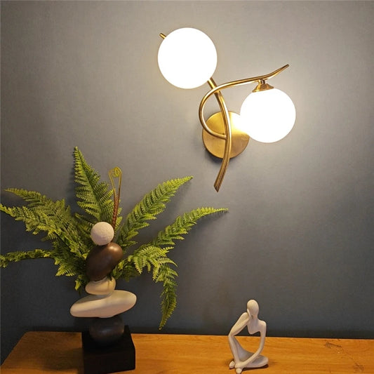 Two-globe wall sconce with frosted glass globes on a gold-color metal branch above a fern and a figurine.