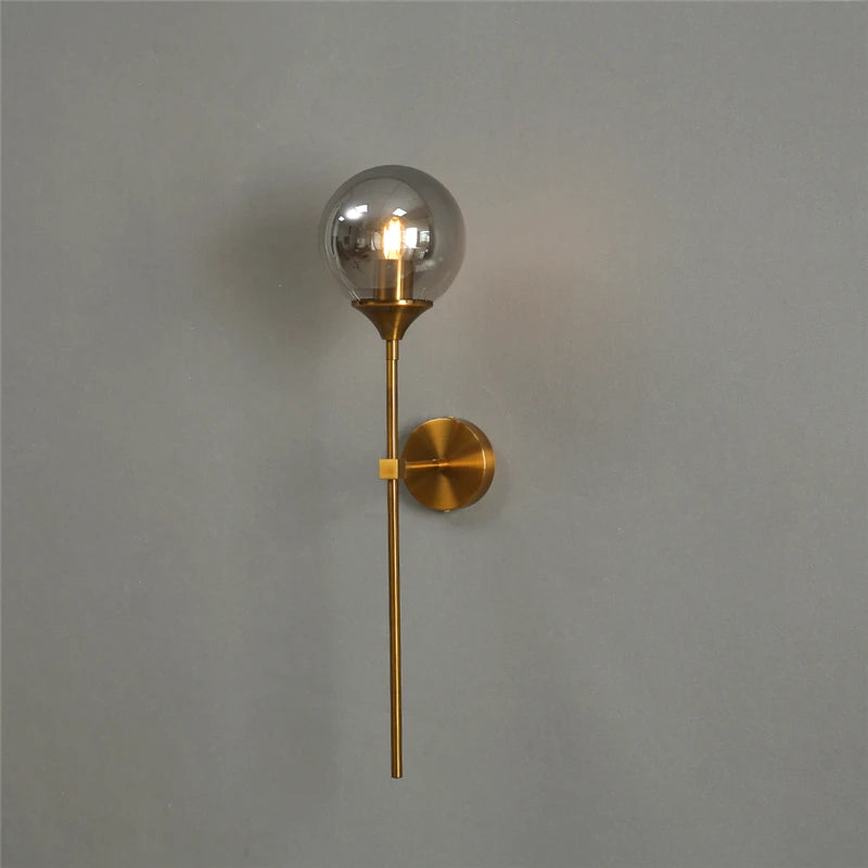 Smoke gray glass globe wall sconce on a gold-color metal stem, lit, seen from the side.