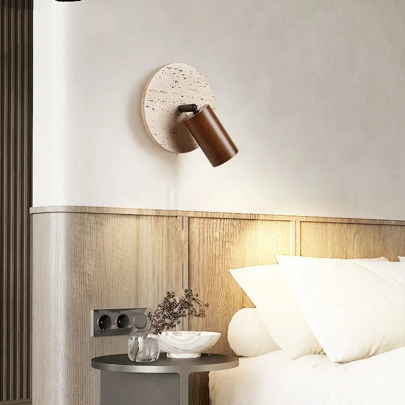 Travertine disc wall sconce with a dark wood spotlight beside a bed