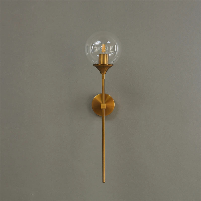 Clear glass globe wall sconce on a long gold-color metal stem, unlit, on a gray wall.