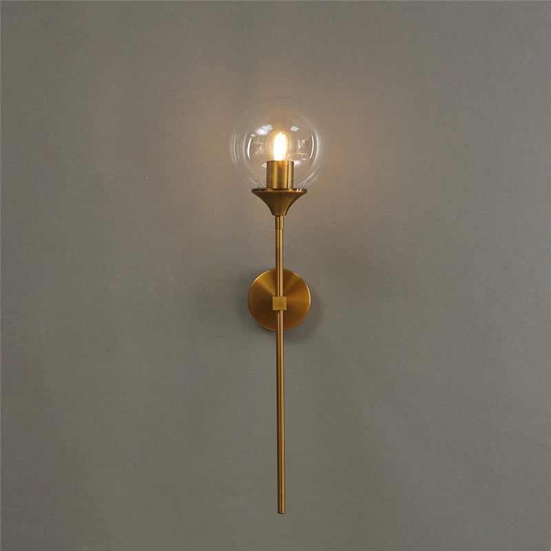 Glass globe wall sconce with a clear globe on a long gold-color metal stem, lit, on a gray wall.