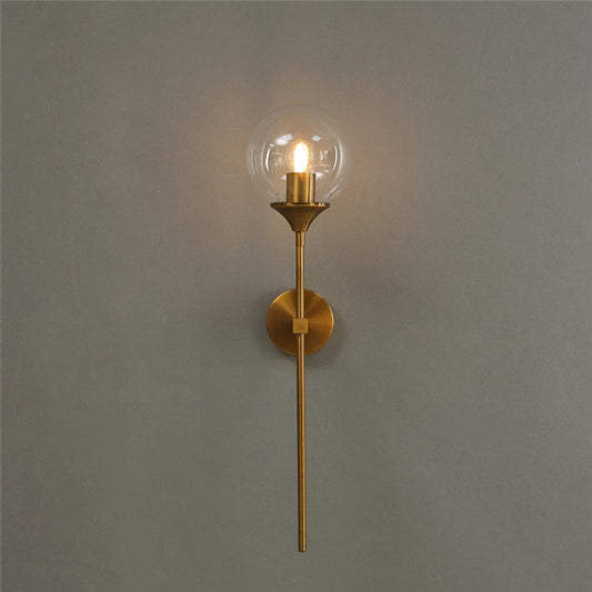 Glass globe wall sconce with a clear globe on a long gold-color metal stem, lit, on a gray wall.