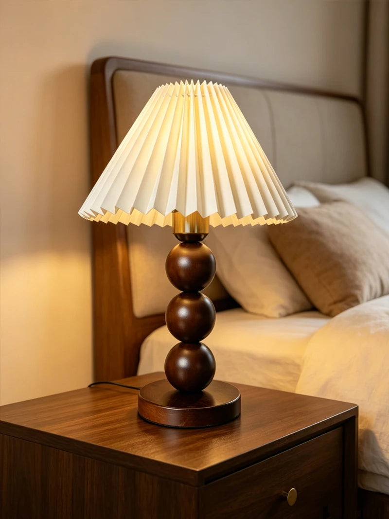 Walnut-color stacked-sphere table lamp with a white pleated shade, lit, on a dark wood nightstand.