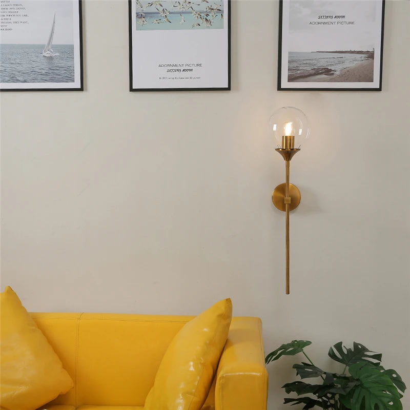 Clear glass globe wall sconce on a gold-color metal stem, lit, on a white wall above a yellow sofa.