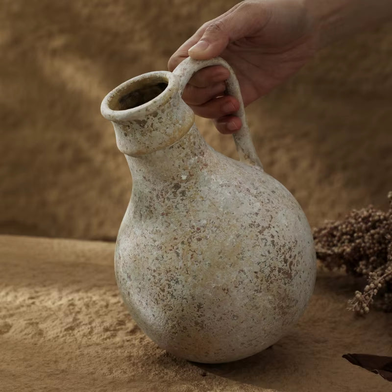 Round rustic ceramic jug vase tilted by its handle, showing the weathered grey finish.