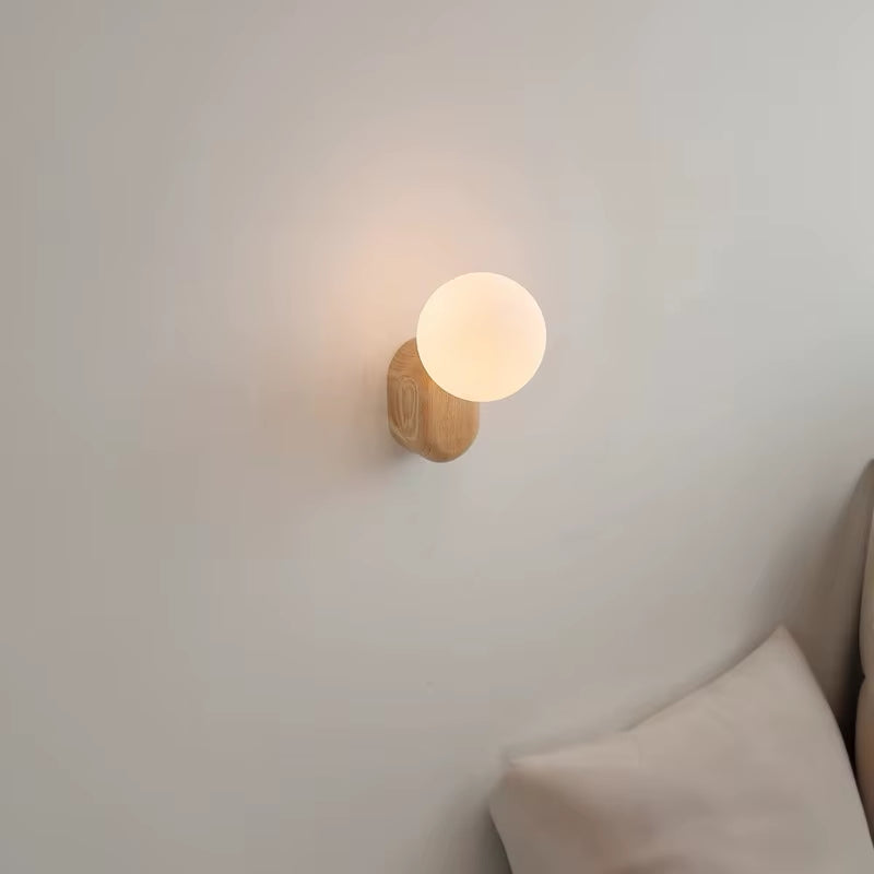 Wall sconce with a white glass globe on a light wood base, lit, seen from the side above a cushion.