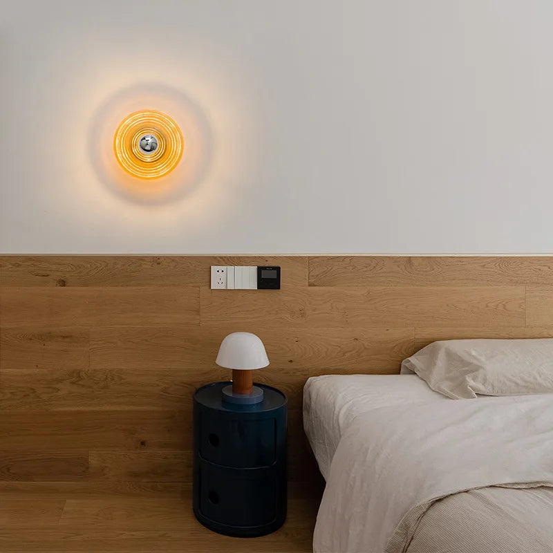 Amber ripple glass disc wall sconce glowing on a white wall above a wood headboard and a bed.