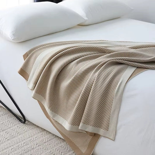 Khaki herringbone knit throw blanket with a cream border draped over the end of a bed.