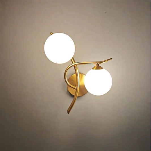 Two-globe wall sconce with frosted glass globes on a curved gold-color metal branch, lit, on a gray wall.