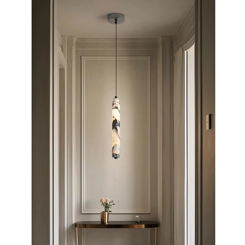 Slim marble cylinder pendant light, lit, hanging from a round ceiling canopy in a hallway above a console table.