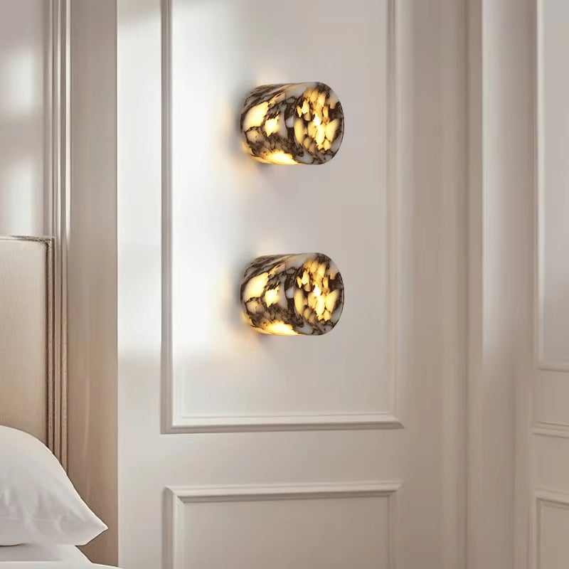 Two marble cylinder wall sconces, lit, one above the other on a panelled wall beside a bed.